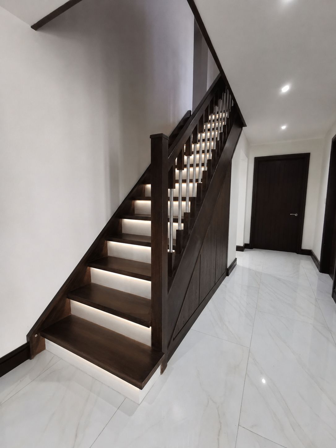 Dark walnut under stairs storage matching a walnut staircase with LED lit treads, beside a marble tiled hallway